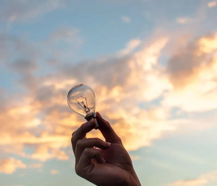 A hand holding a lightbulb against a dawn-filled sky