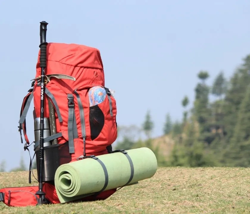 A red backpack. There is a green rolled-up sleeping mat attached to the backpack. 