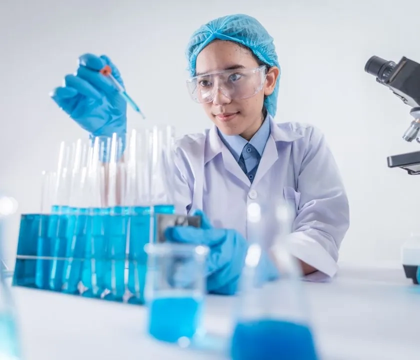 A scientist dripping blue liquid into a test tube using a pipette. She is in a lab.