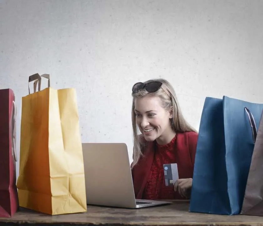 A woman holding a credit card in front of a laptop, surrounded by shopping bags